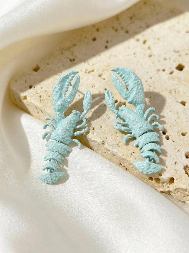 Pair of mint blue lobster-shaped drop earrings resting on a travertine stone tile against white satin fabric