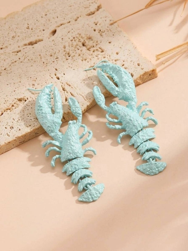 Two mint blue lobster earrings displayed flat on a sandy travertine surface with dried grass in background