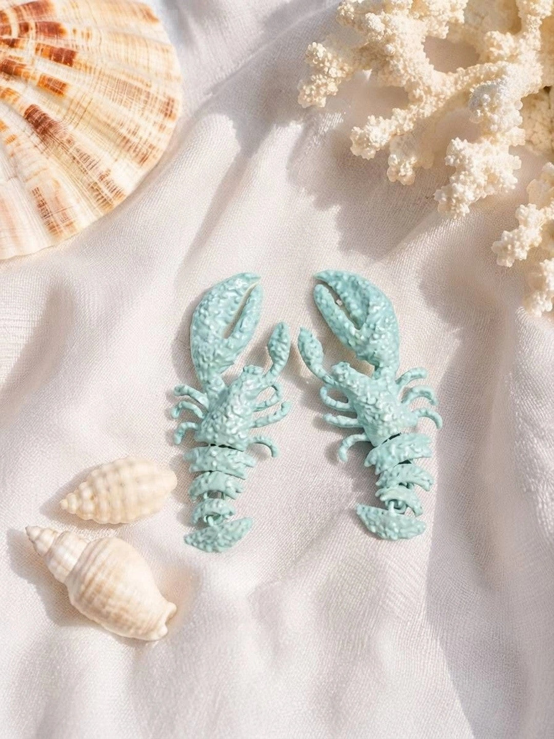 Mint blue lobster drop earrings placed on white fabric surrounded by seashells and white coral pieces