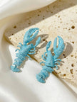 Two sky blue lobster-shaped drop earrings resting on a travertine stone tile against white satin