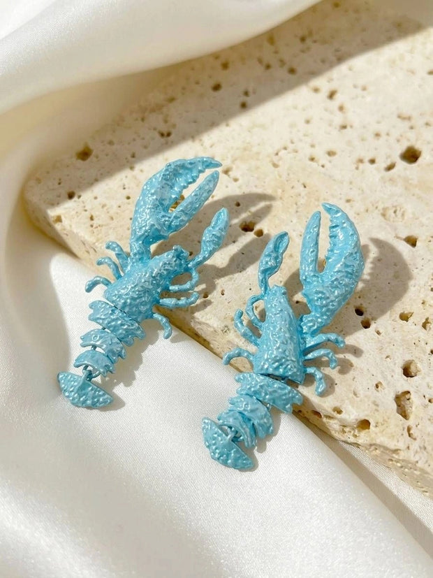 Two sky blue lobster-shaped drop earrings resting on a travertine stone tile against white satin