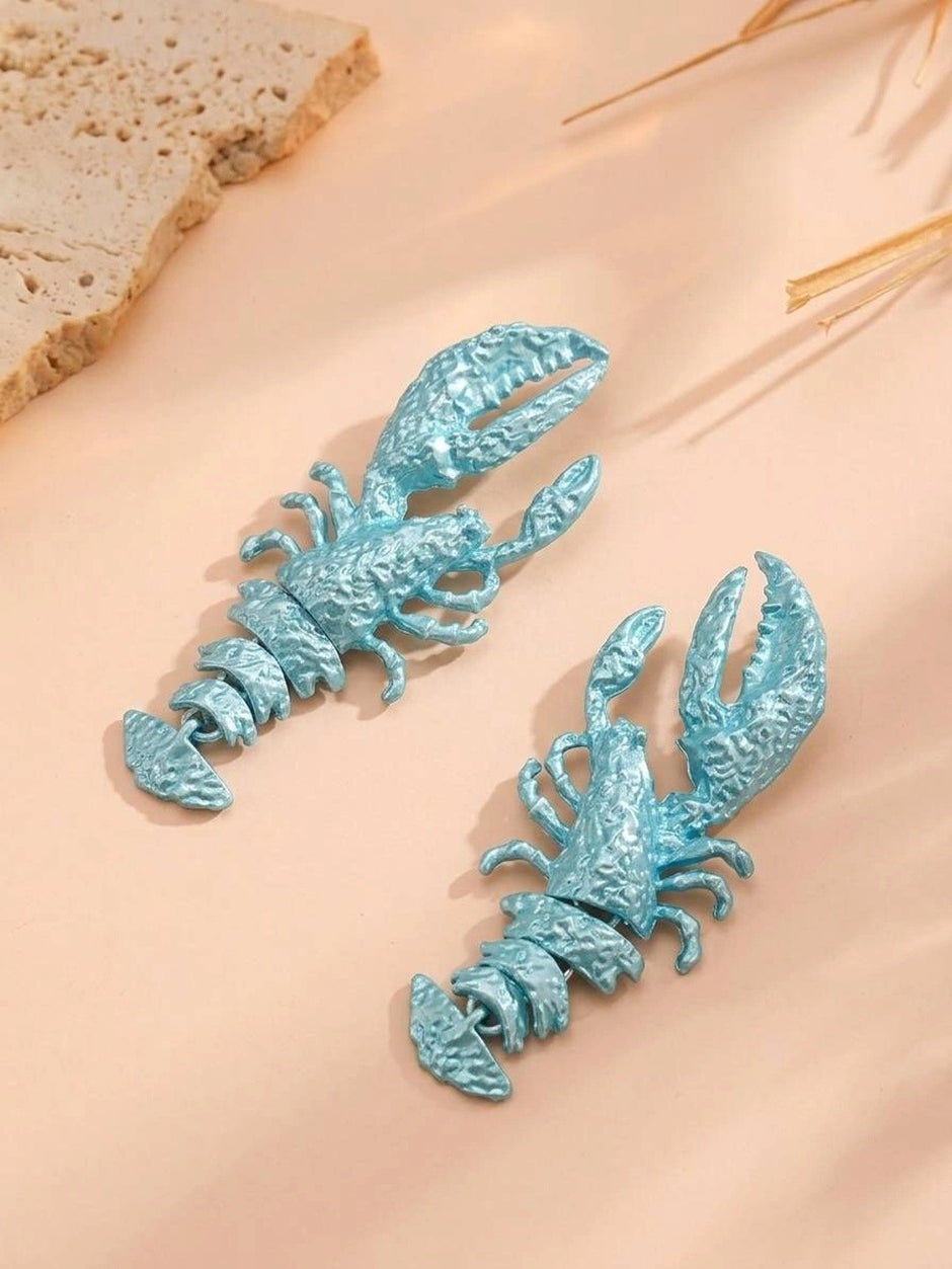 Pair of pearlescent sky blue lobster earrings laid flat on a warm peach surface with dried grass
