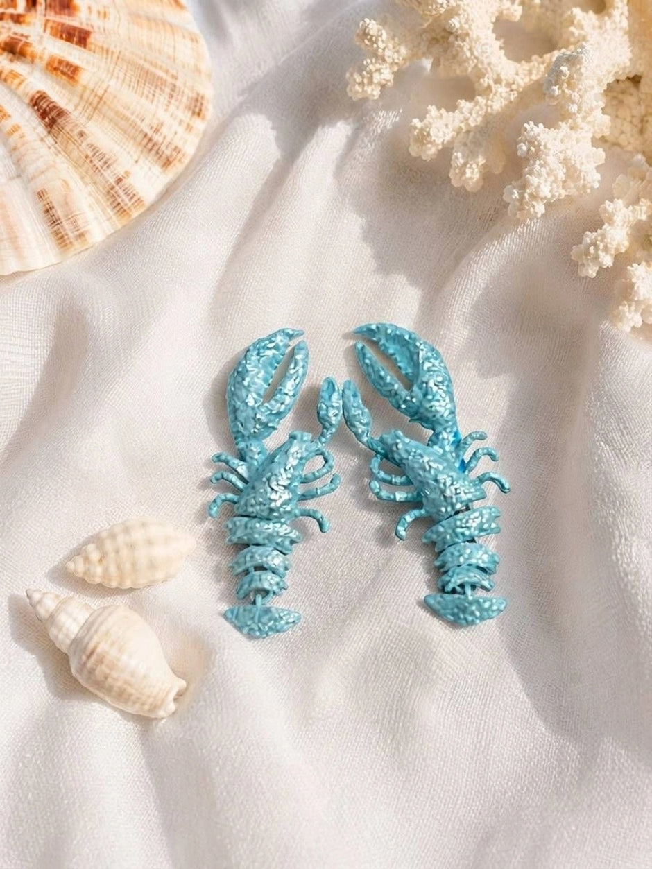 Sky blue lobster earring pair displayed on white fabric beside seashells and white coral