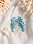 Sky blue lobster earring pair displayed on white fabric beside seashells and white coral