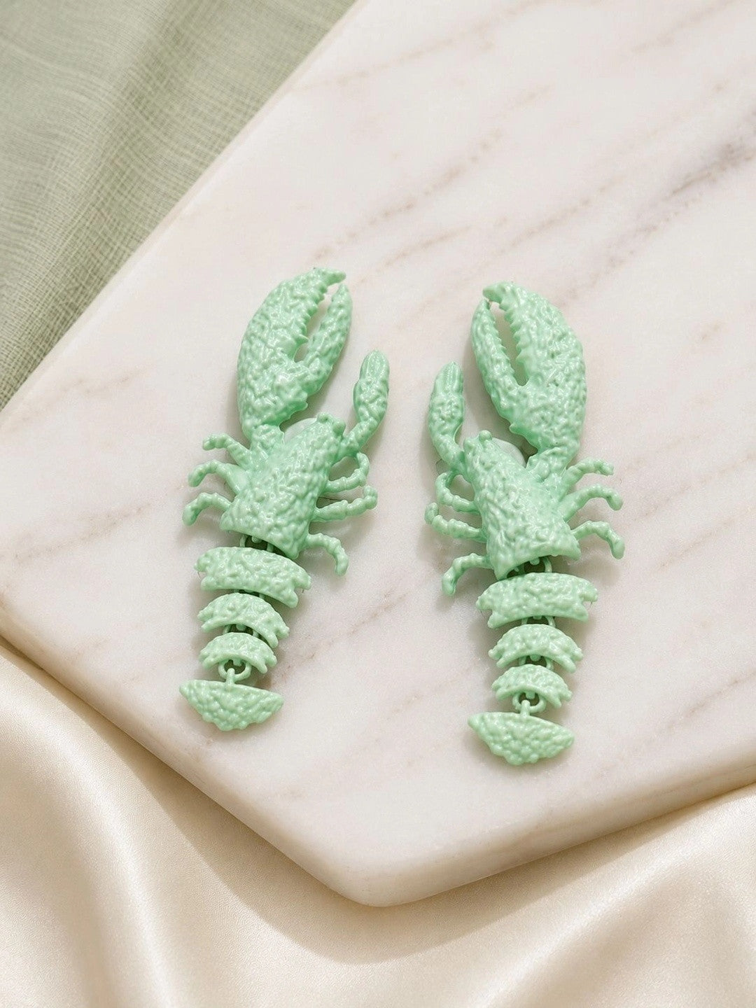 Pair of mint green lobster drop earrings laid flat on a white marble surface