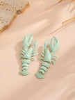 Two mint green lobster earrings displayed on a peach surface with dried grass