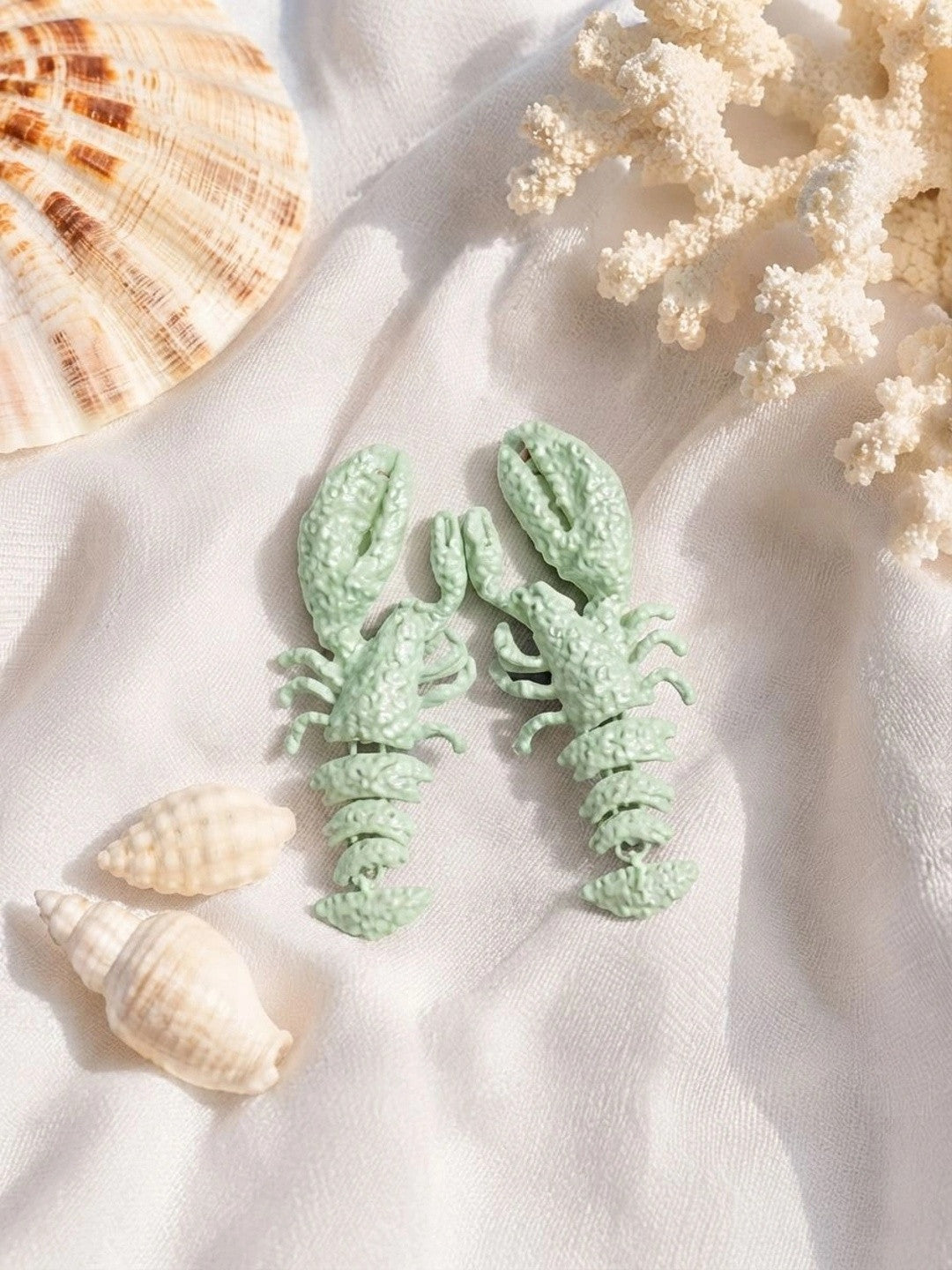 Mint green lobster earrings styled on white cloth alongside seashells and coral