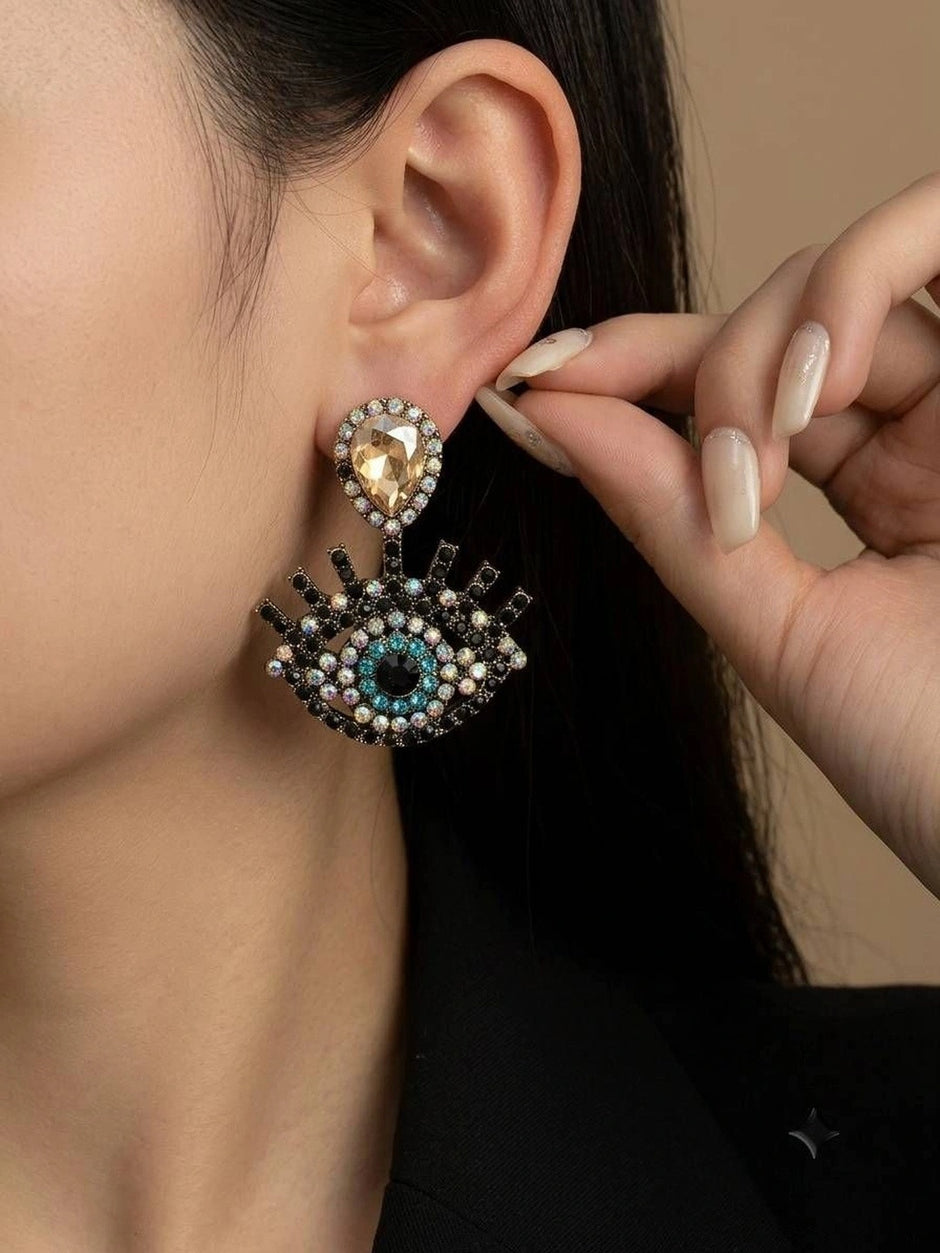 Model wearing gold-tone evil eye crystal drop earring with champagne teardrop stud and black rhinestone lashes