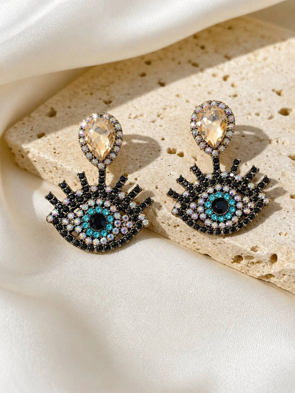 Two evil eye crystal earrings on textured stone surface highlighting teal and iridescent AB stones