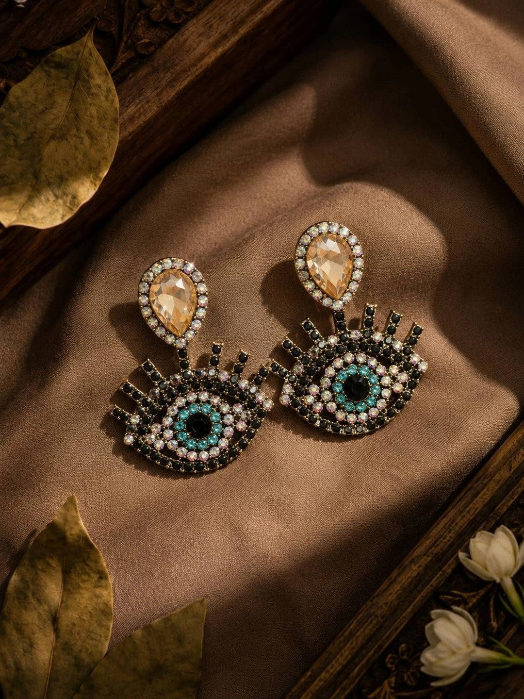 Evil eye rhinestone drop earrings displayed on brown satin with dried leaves and white flowers