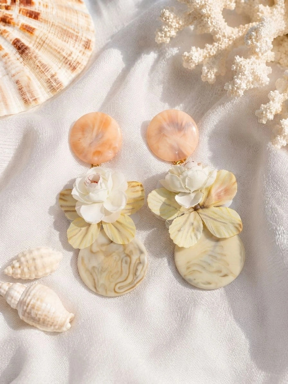 Pair of peach resin stud and cream swirl disc drop earrings with yellow fabric butterfly and white floral detail, styled on white fabric with seashells
