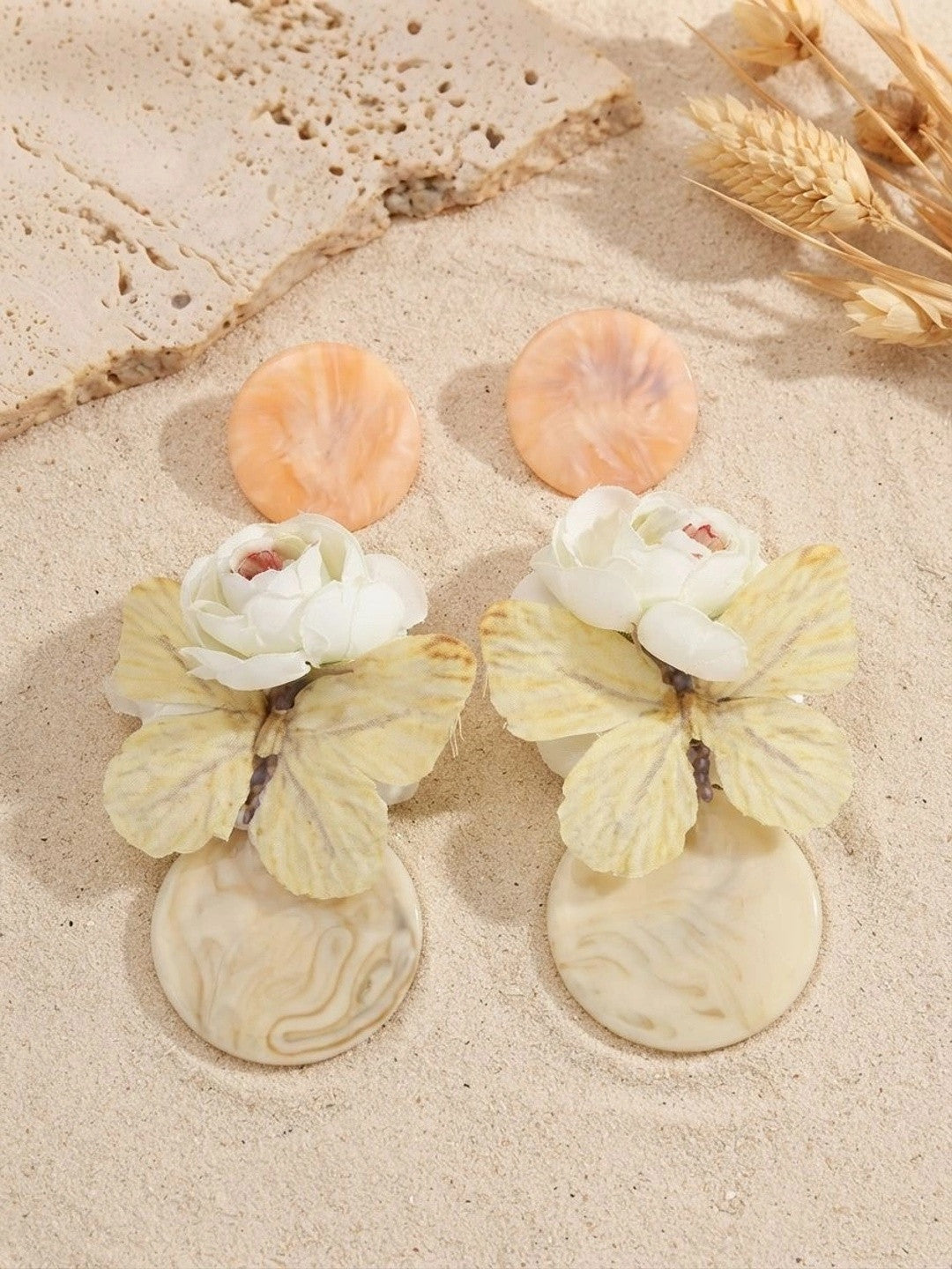 Peach and cream resin butterfly drop earring pair displayed on sand with dried wheat stalks in warm sunlight