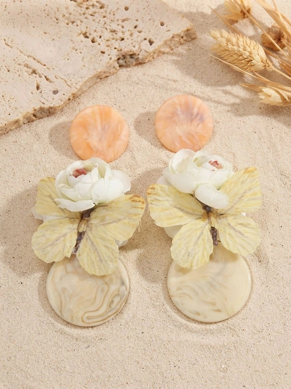 Peach and cream resin butterfly drop earring pair displayed on sand with dried wheat stalks in warm sunlight