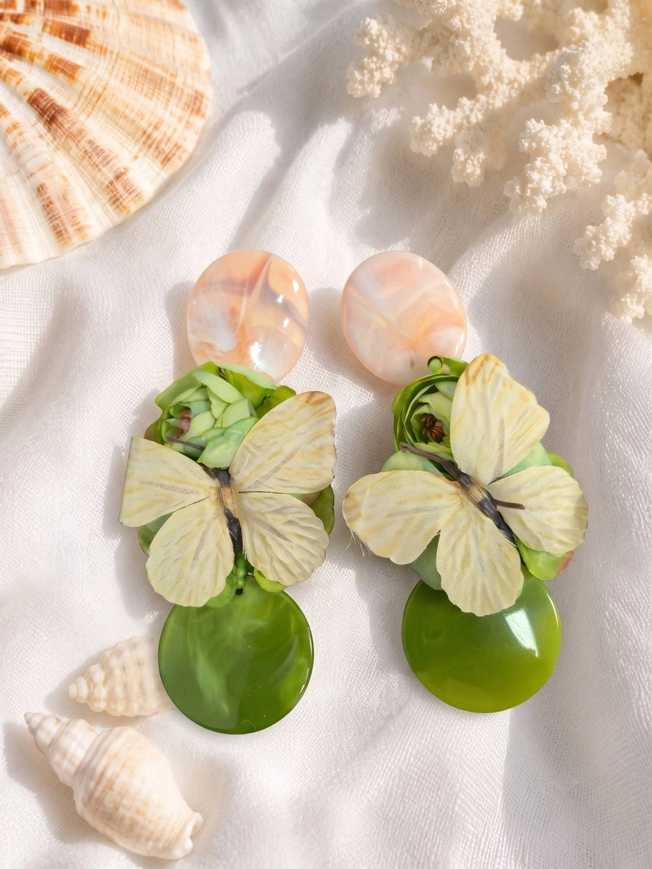 Pair of green resin and butterfly drop earrings laid flat on white fabric with seashells
