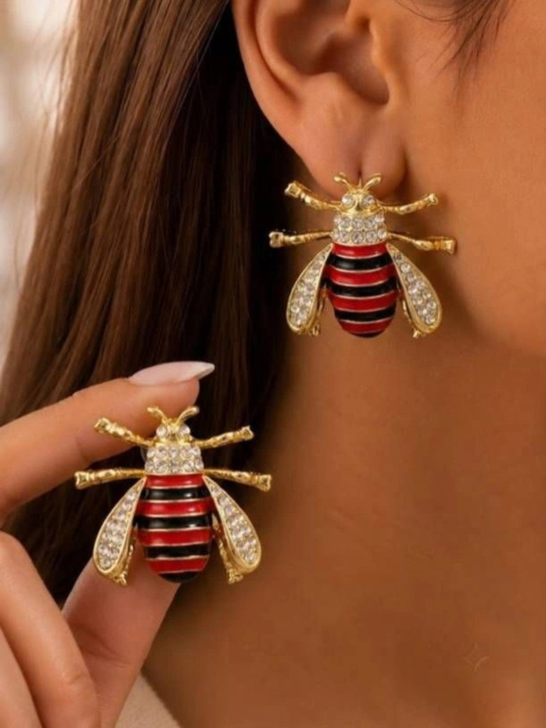 Model wearing gold bee stud earrings with red black enamel and crystal wings on ear, holding second earring