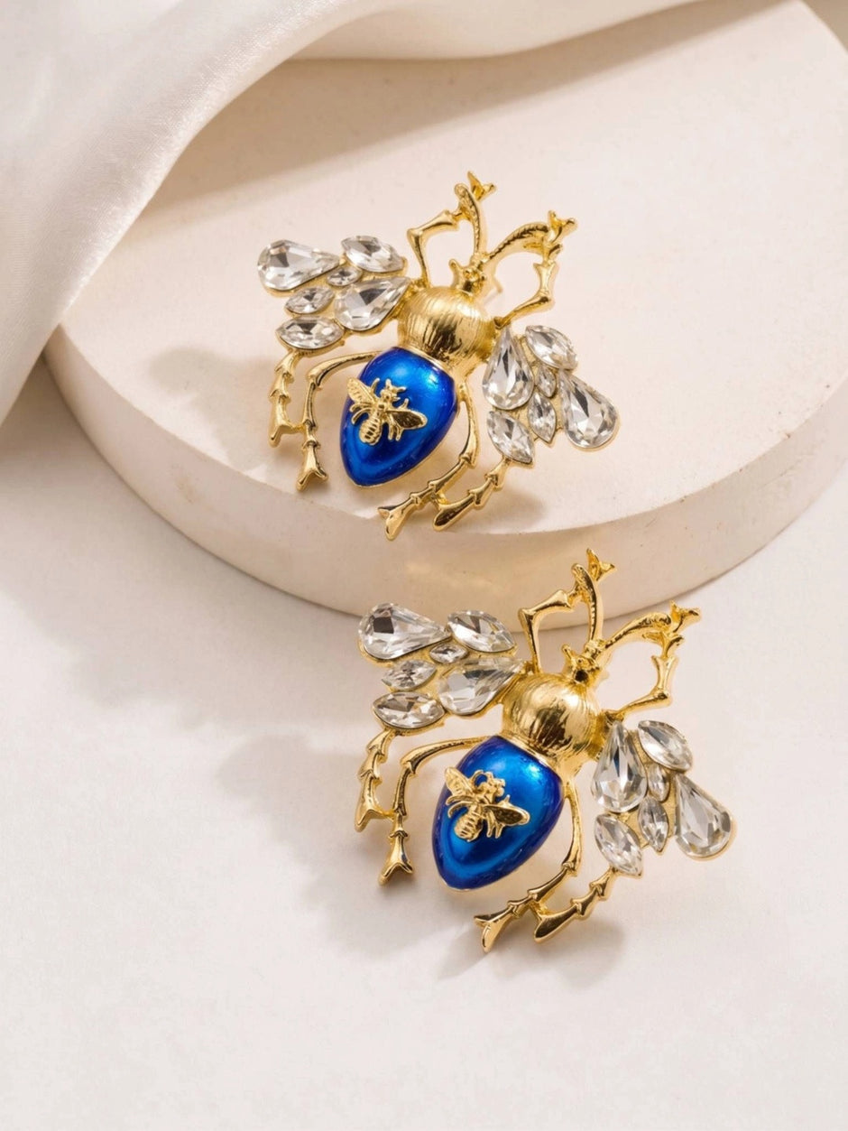 Pair of blue enamel beetle earrings with crystal wings resting on a white stone surface