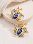 Pair of blue enamel beetle earrings with crystal wings resting on a white stone surface