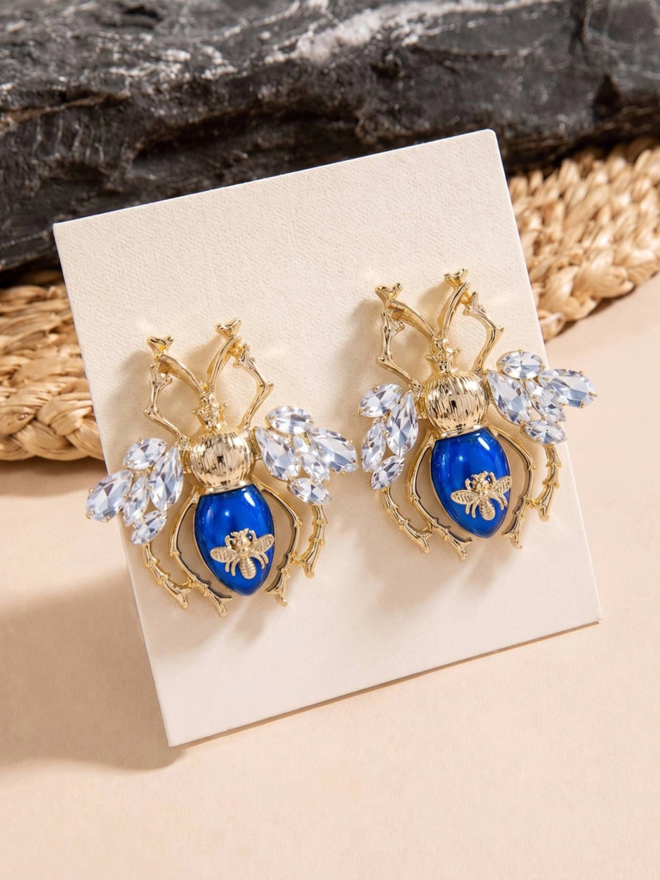 Two gold and blue beetle stud earrings displayed upright on a white jewellery card