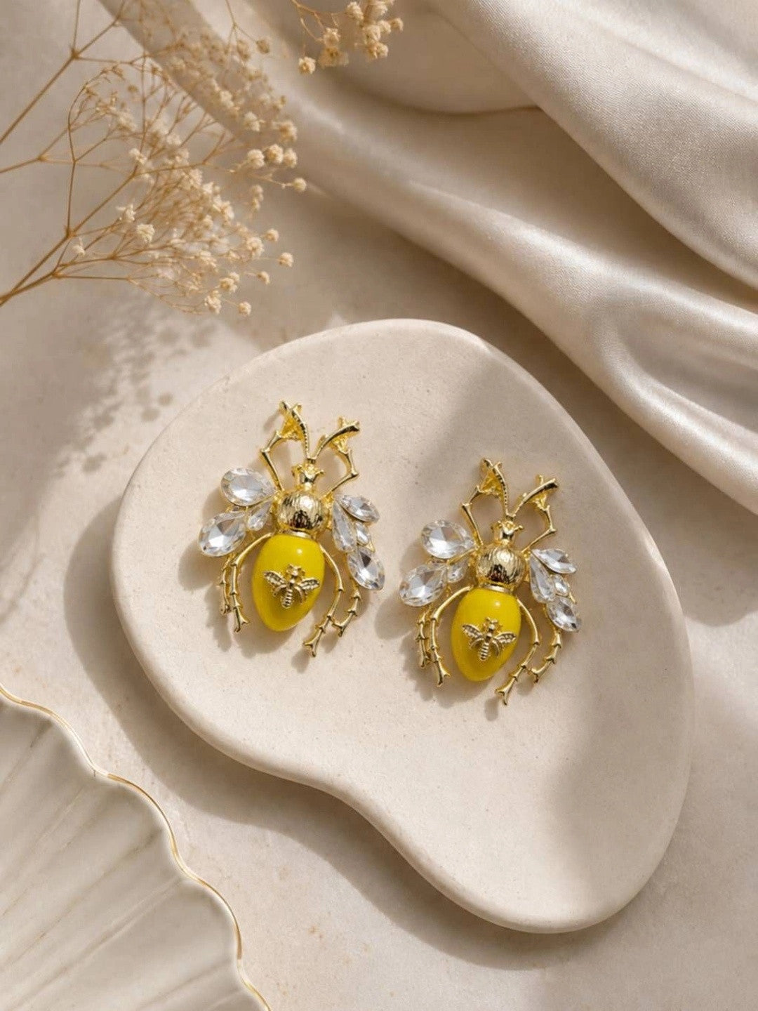 Pair of gold bee stud earrings with crystal wings placed on a cream ceramic dish with dried flowers