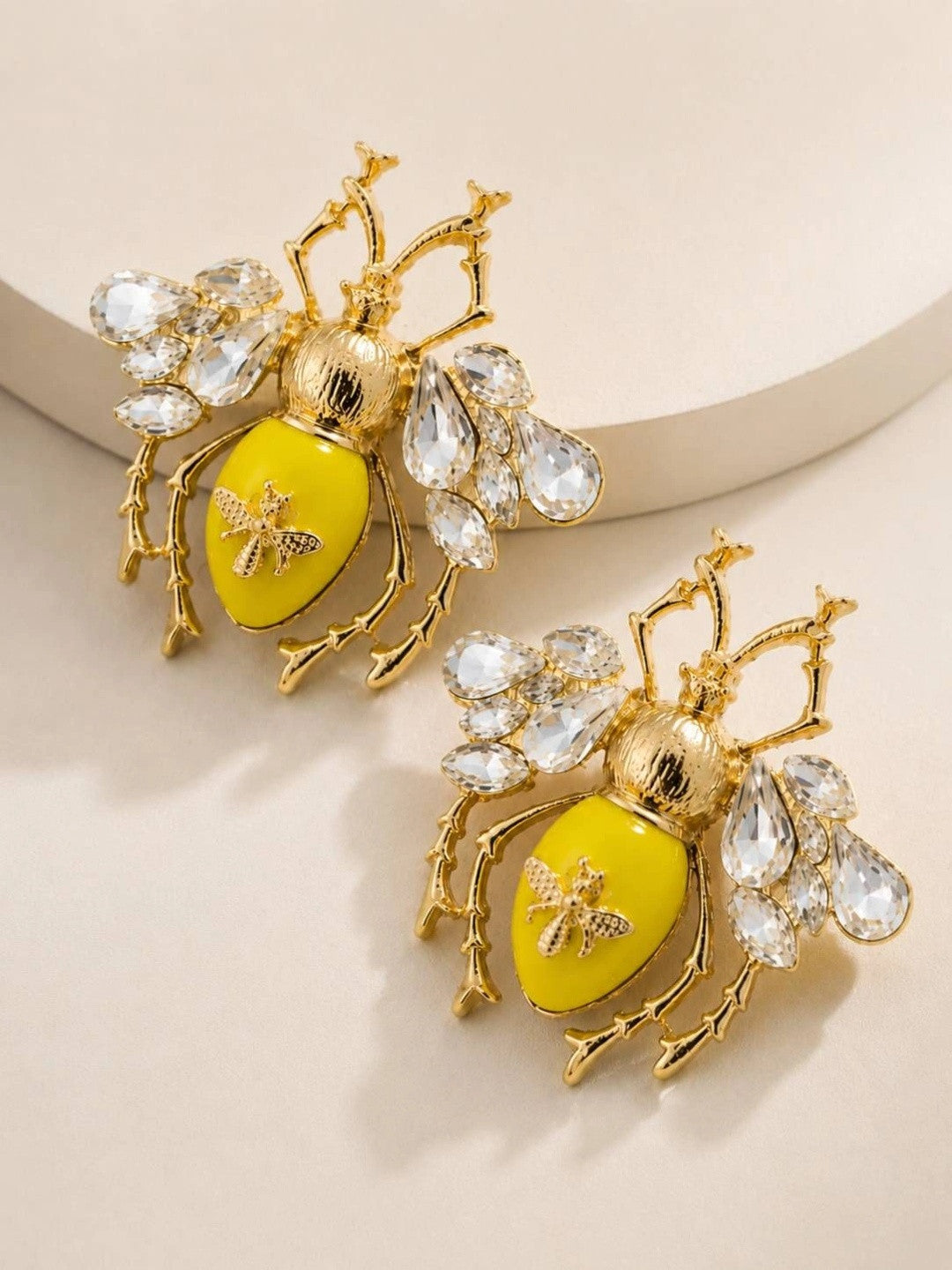 Two gold-plated bee earrings with yellow enamel and faceted crystal wings resting on a beige surface