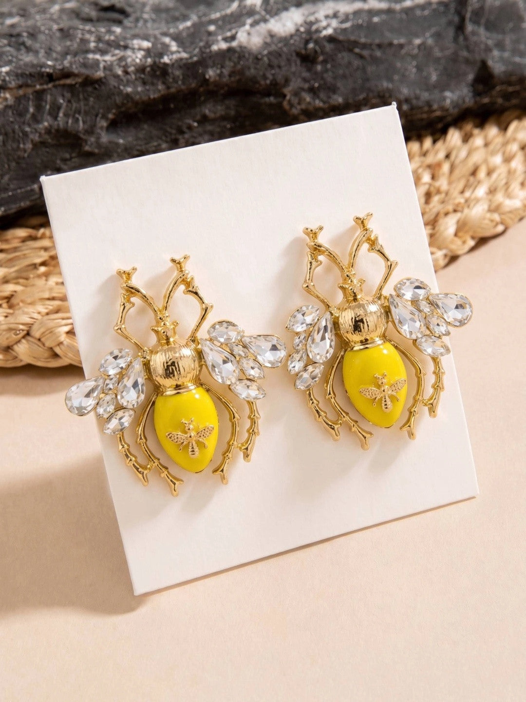 Gold bee stud earrings with yellow enamel body displayed on a white jewellery card against a textured background