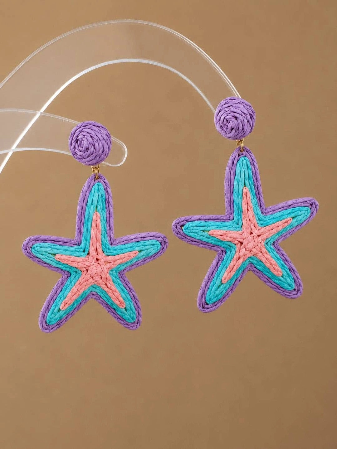 Pair of raffia woven starfish drop earrings in purple, turquoise and pink displayed on a clear acrylic stand against a warm beige background
