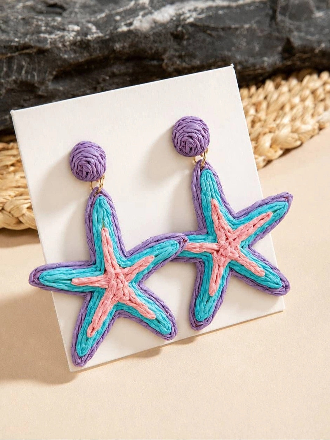 Close-up of raffia starfish earrings with purple ball studs resting on a white earring card with a woven mat backdrop