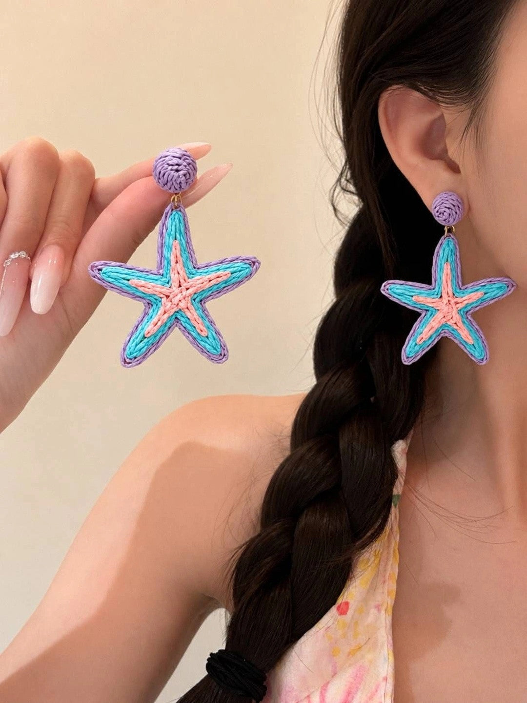 Model wearing and holding raffia starfish drop earrings, showing scale and fit against her ear with a braided hair backdrop
