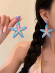 Model wearing and holding raffia starfish drop earrings, showing scale and fit against her ear with a braided hair backdrop