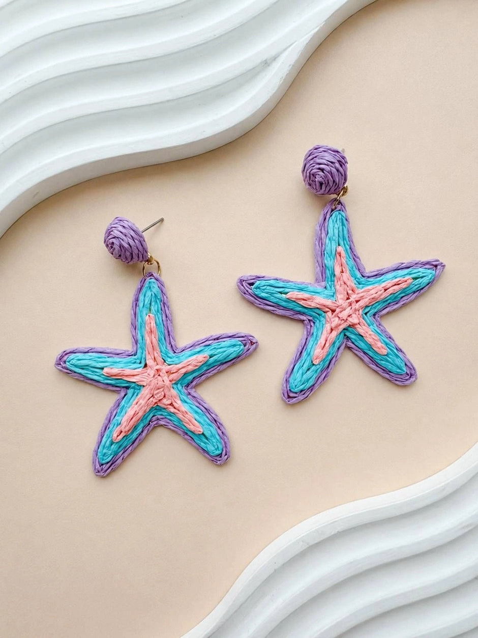 Both raffia starfish earrings laid flat on a peach surface beside a white sculptural prop showing full design detail