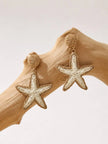 Both woven beige and white starfish drop earrings hanging from a driftwood branch against white background
