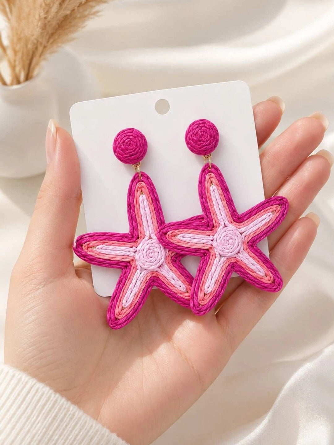 Both pink raffia starfish earrings displayed on a white card held in a woman's palm