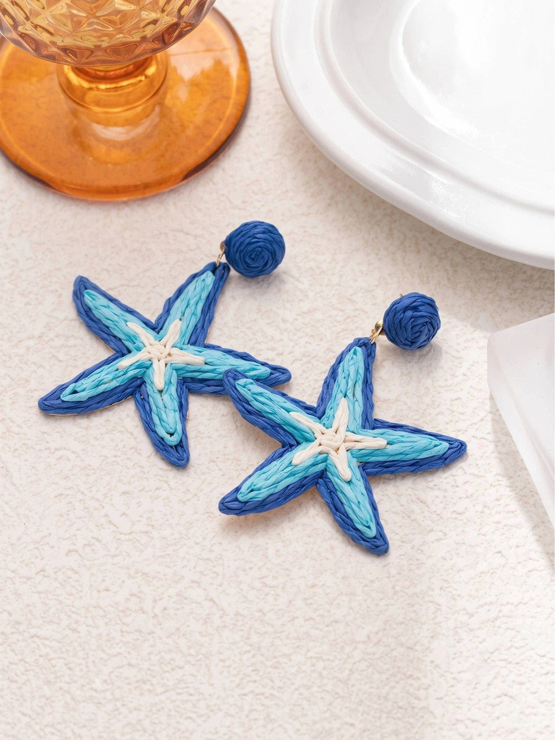Two raffia starfish earrings with cobalt and sky blue cord laid flat on a white textured surface