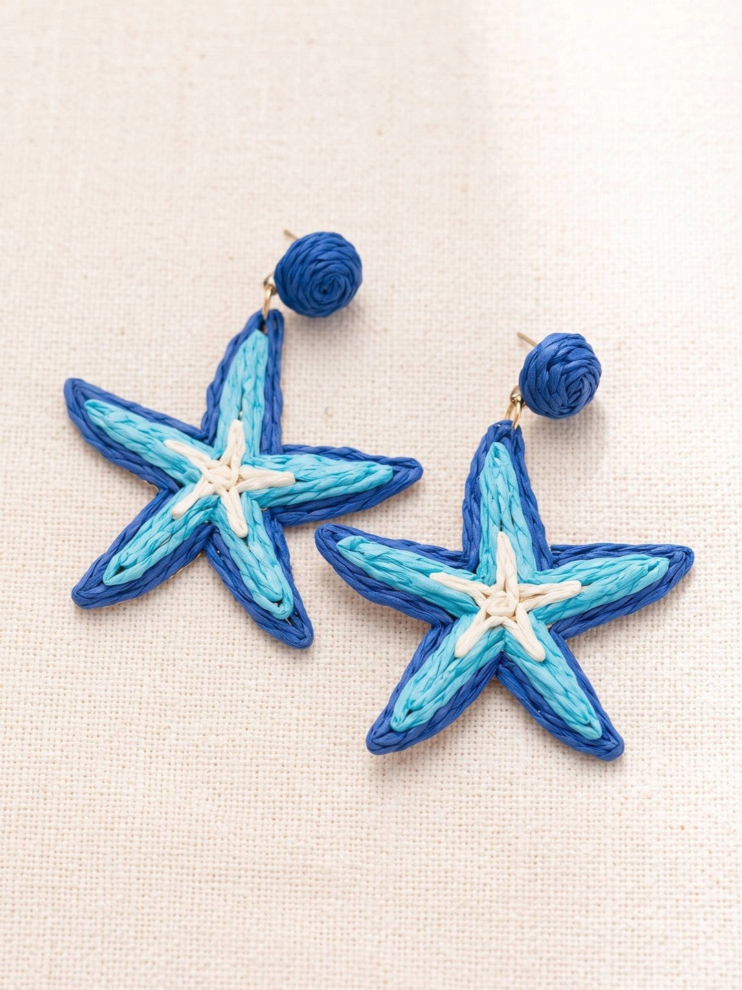 Close-up flat lay of raffia starfish earring pair showing cobalt blue border and cream centre detail on linen
