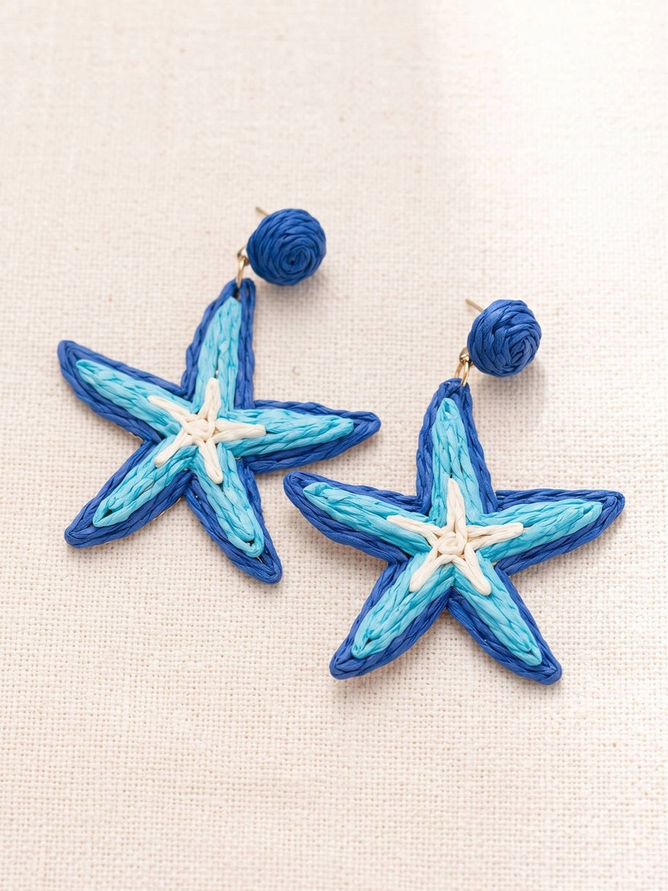 Close-up flat lay of raffia starfish earring pair showing cobalt blue border and cream centre detail on linen