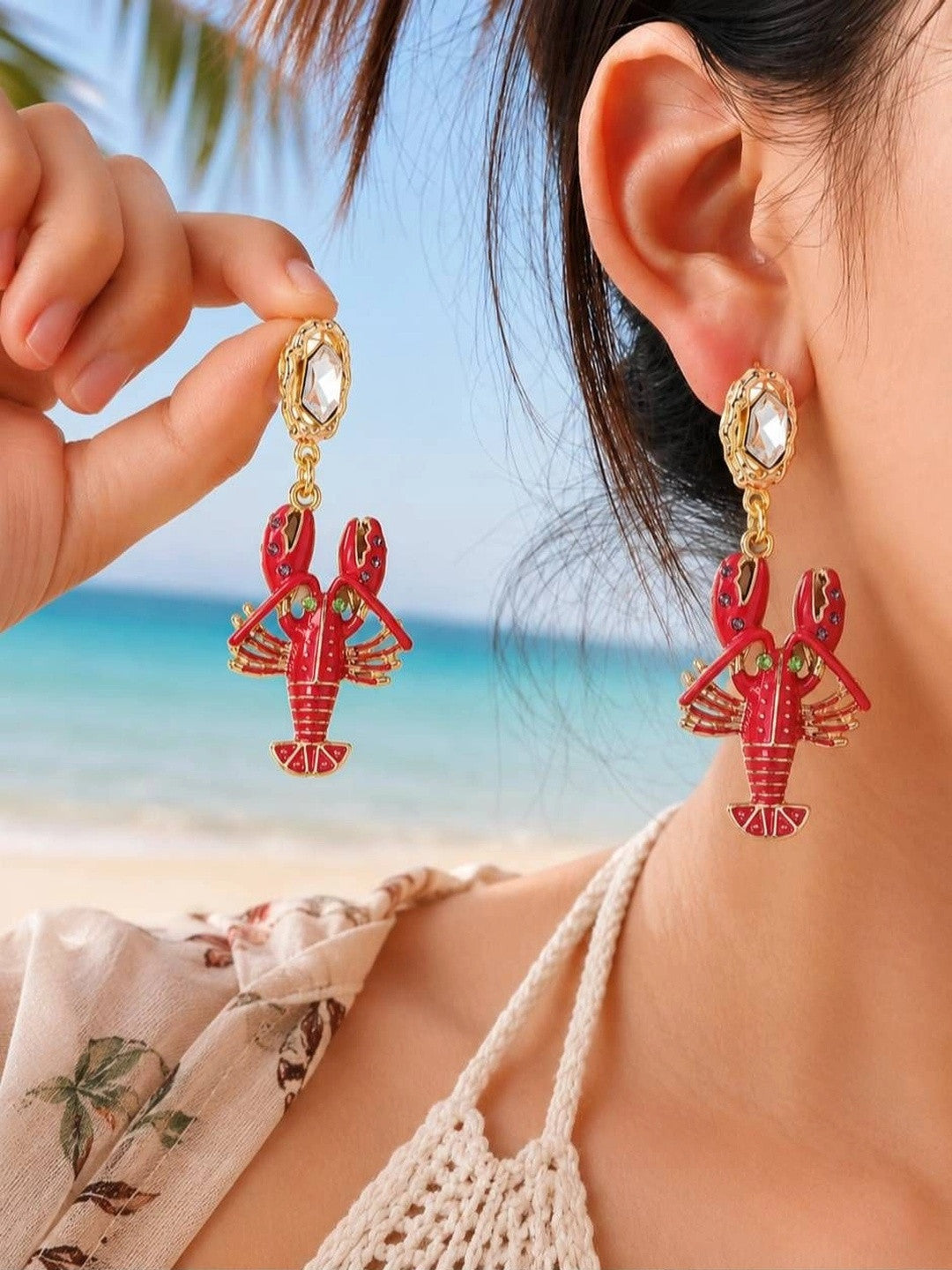 Model wearing fuchsia lobster drop earrings at a beach, holding one earring up to show detail