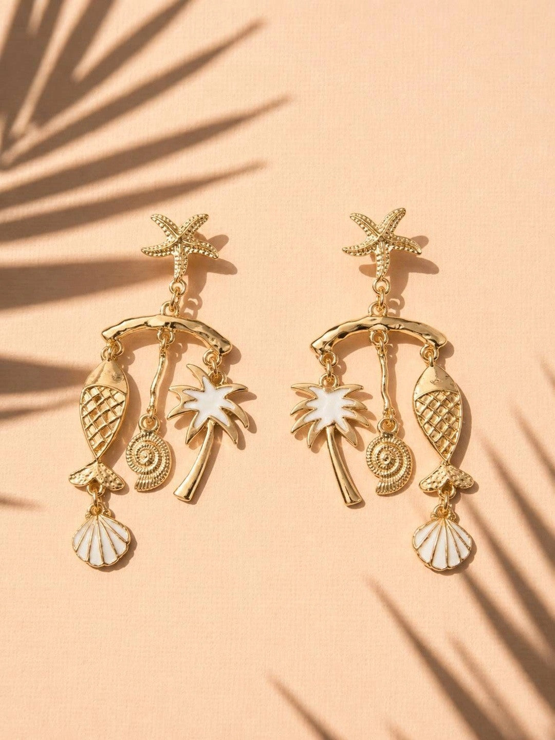 Gold-plated beach charm drop earrings with white enamel accents displayed on peach surface with palm leaf shadow