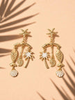 Gold-plated beach charm drop earrings with white enamel accents displayed on peach surface with palm leaf shadow