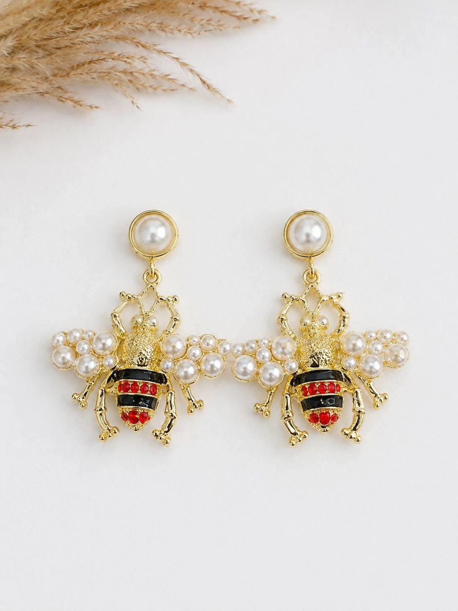 Pair of pearl and gold bee drop earrings laid flat on white surface with dried grass in background