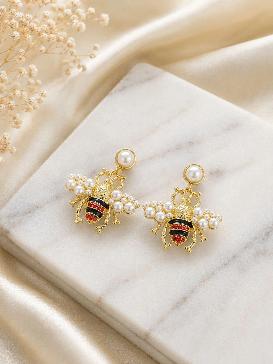 Two gold bee earrings with pearl-studded wings displayed on marble surface with dried white flowers