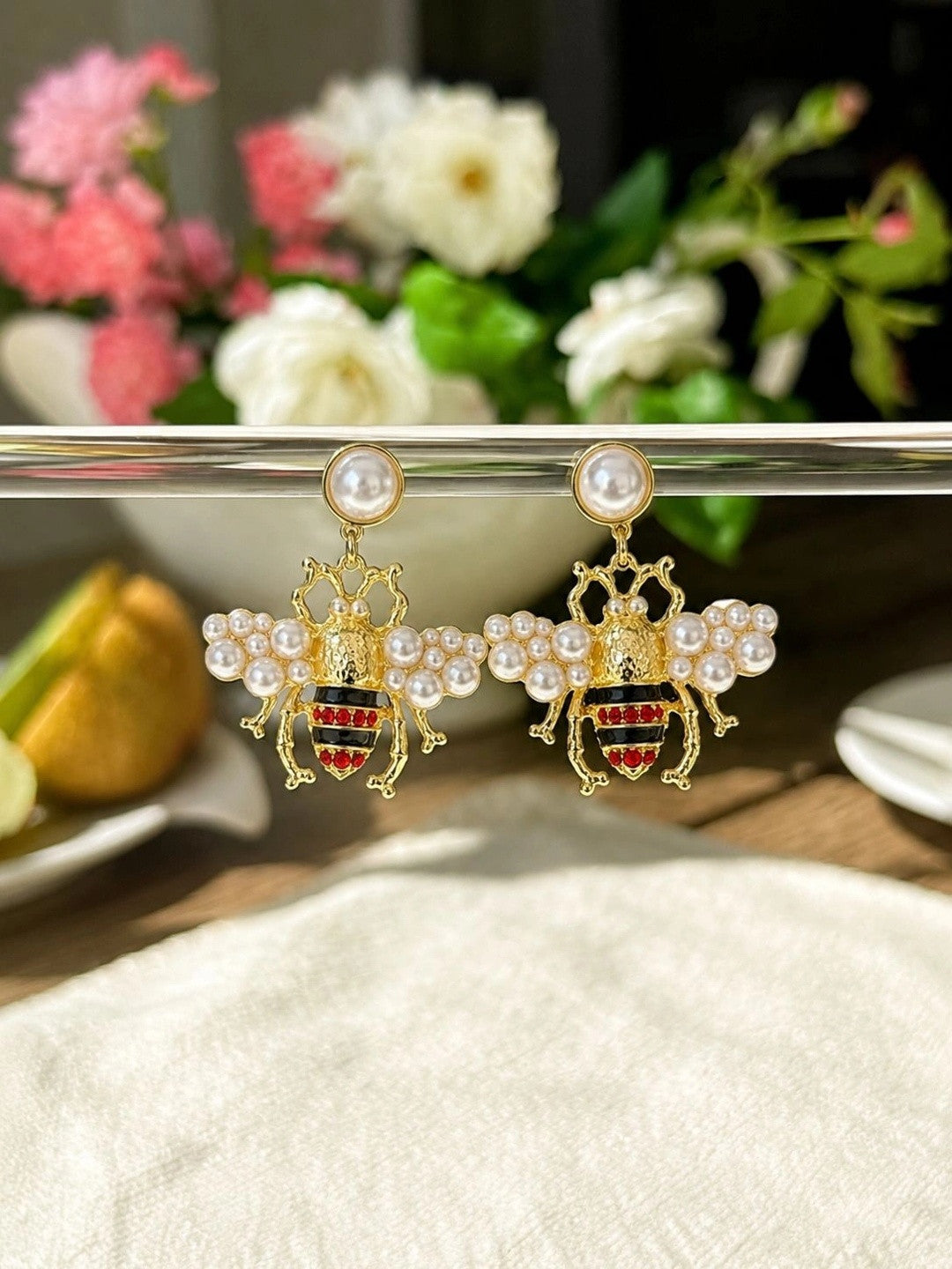 Gold pearl bee earrings hanging from a metal display bar with floral arrangement in background