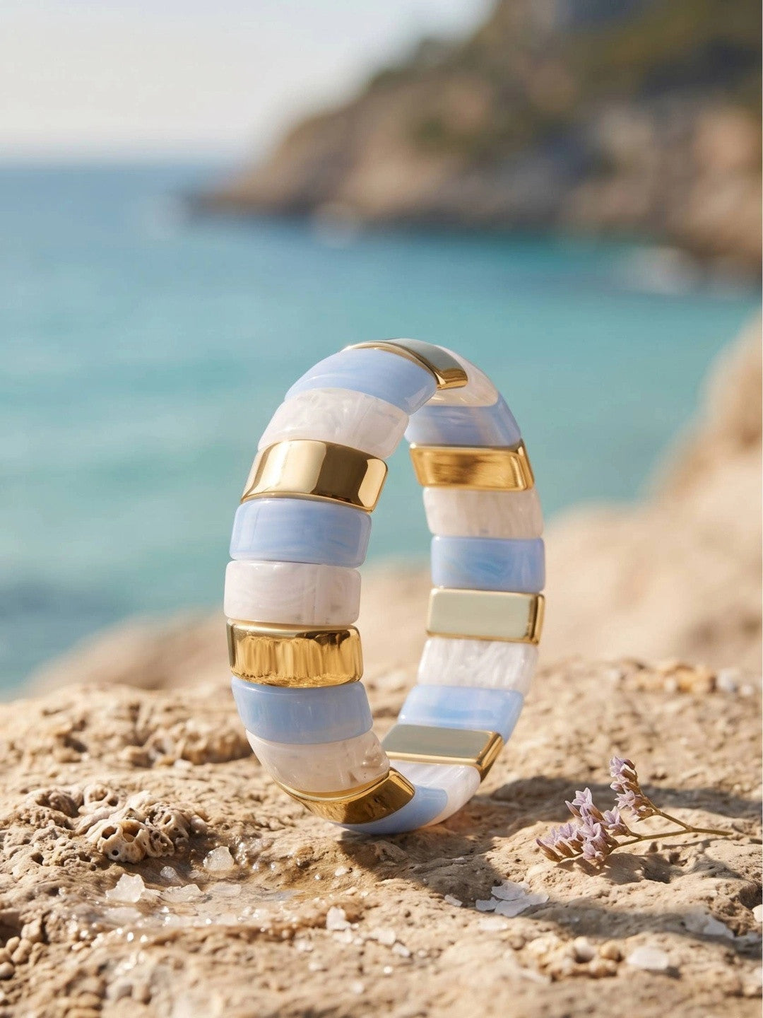 Blue and white resin striped cuff bangle with gold-toned bars resting on a sandy coastal rock with turquoise sea in the background