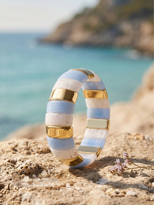 Blue and white resin striped cuff bangle with gold-toned bars resting on a sandy coastal rock with turquoise sea in the background