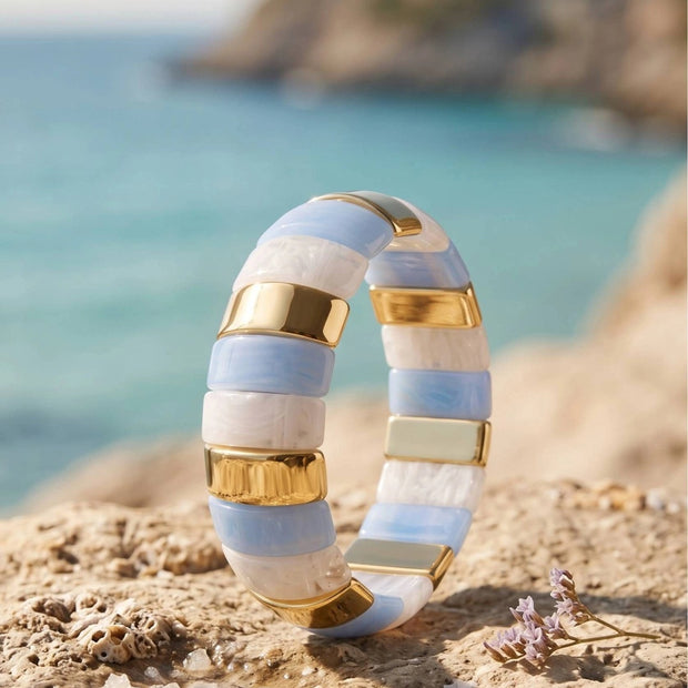 Blue and white resin striped cuff bangle with gold-toned bars resting on a sandy coastal rock with turquoise sea in the background