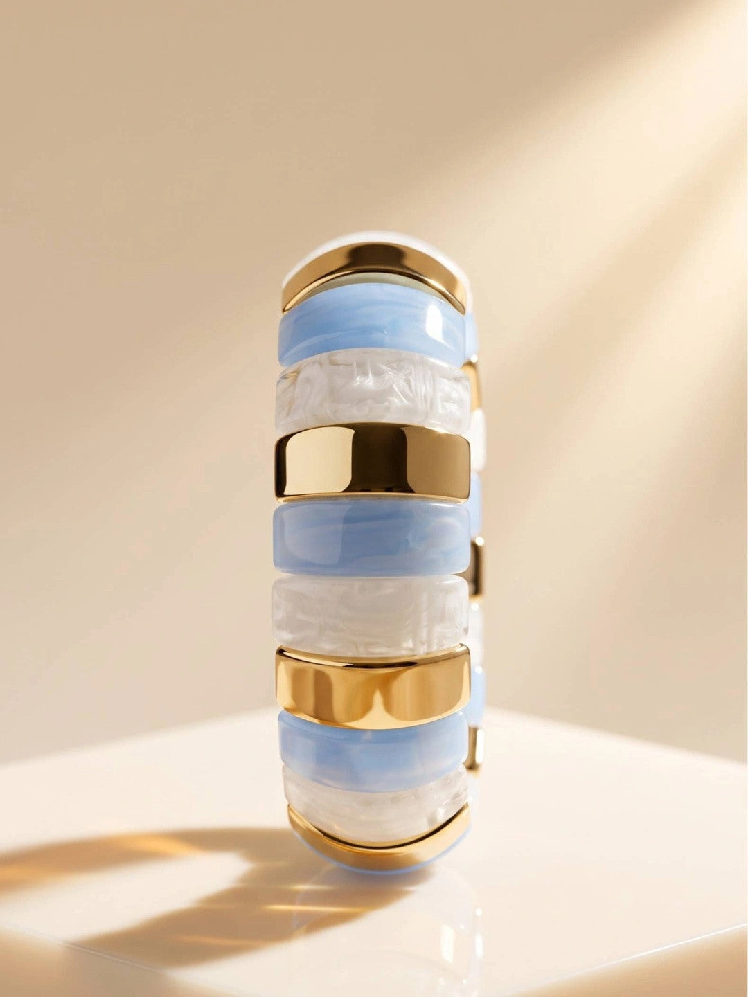 Close-up front view of blue and white resin cuff bangle showing alternating resin segments and polished gold-toned metal spacers on a cream surface
