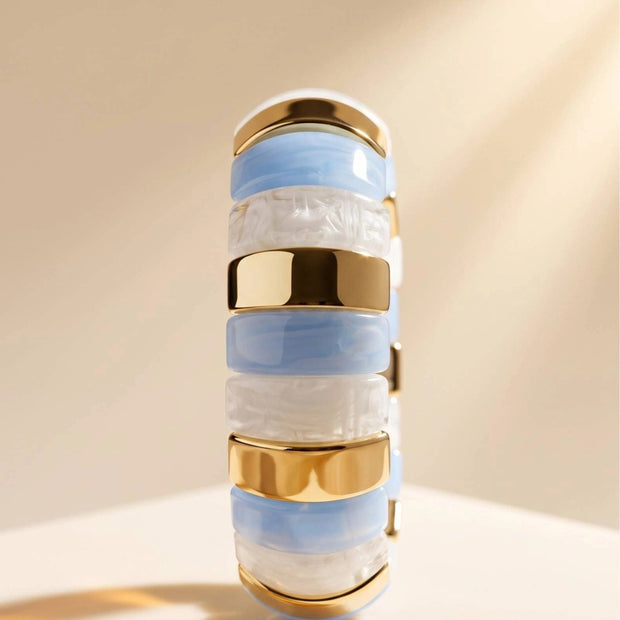 Close-up front view of blue and white resin cuff bangle showing alternating resin segments and polished gold-toned metal spacers on a cream surface