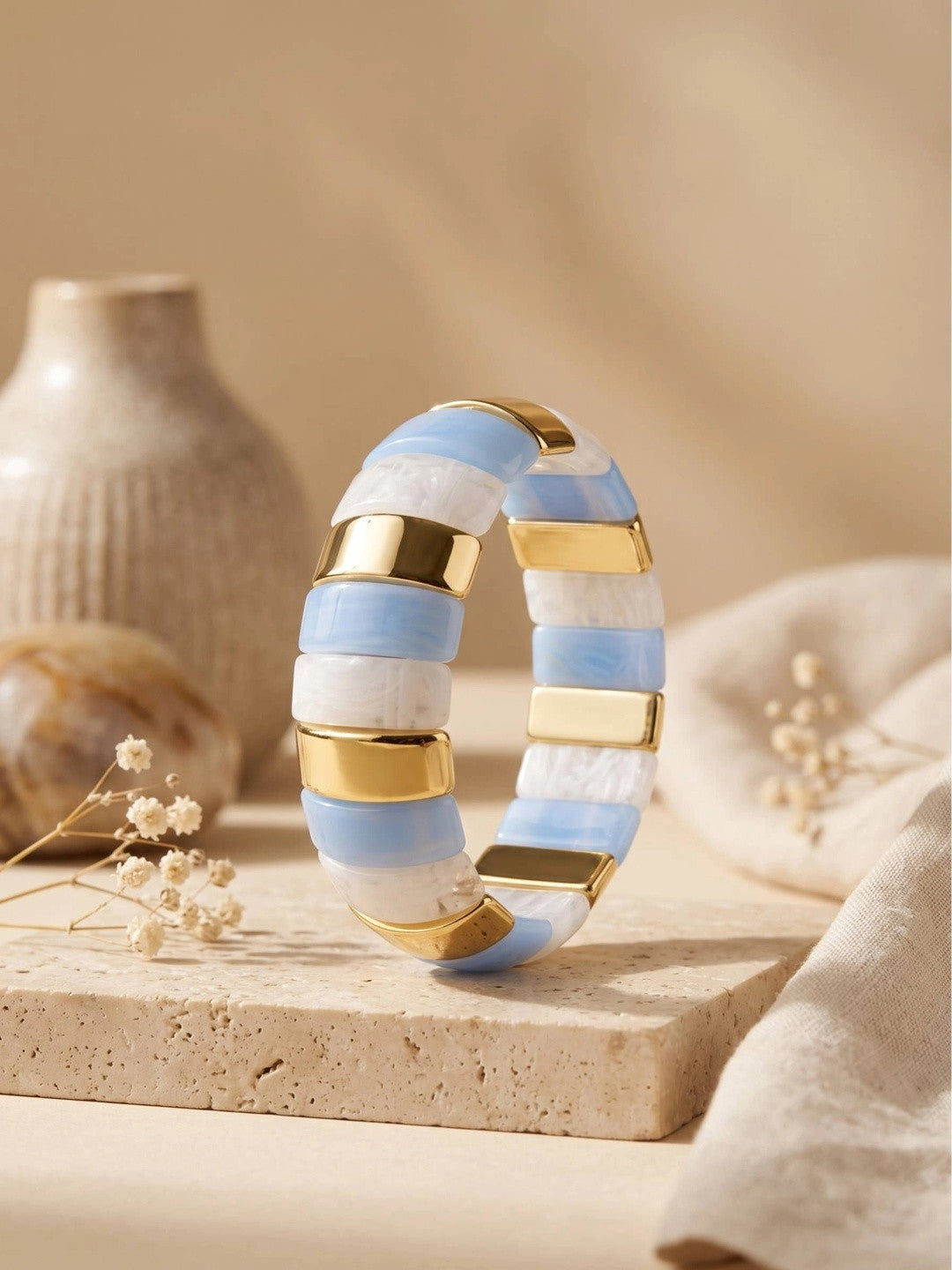 Gold-toned blue resin striped cuff bangle styled on a travertine surface beside dried flowers and a ceramic vase