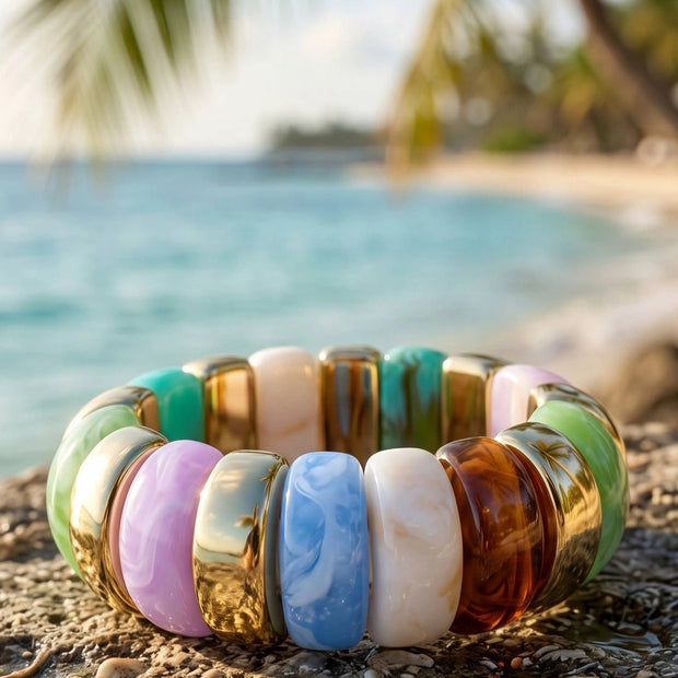 Multicolour resin stretch bracelet with gold-tone spacers resting on a coral rock beside a tropical beach