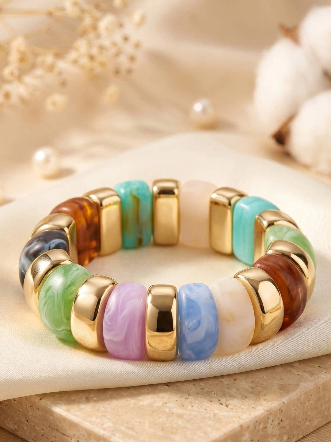 Flat lay of multicolour marbled resin stretch bracelet with gold spacers on a cream fabric surface with pearls