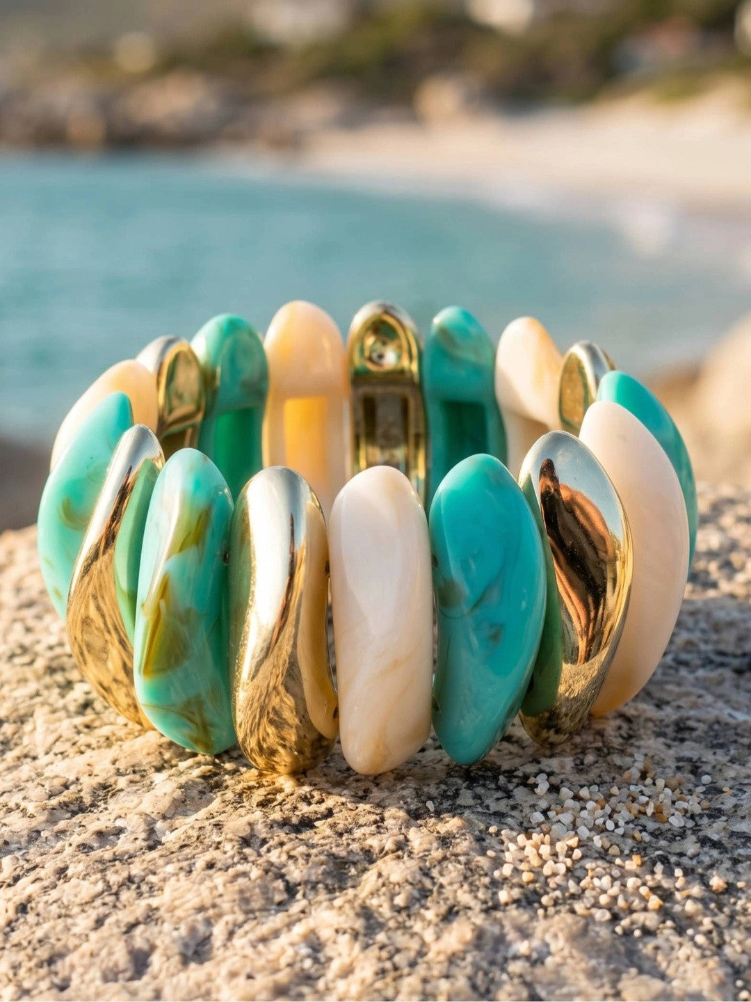 Turquoise marble and gold-tone resin cuff bracelet resting on a sandy rock with blurred sea in background